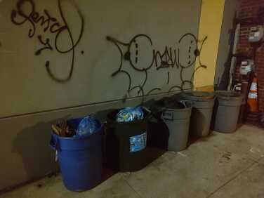 A row of recycling and garbage cans next to a wall with graffiti. 