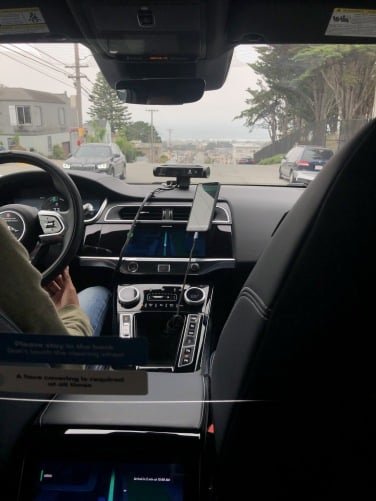 The view from the backseat of a self-driving taxi showing cars parked along the street and oncoming traffic. 